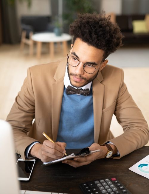 Concentrated young mixed race businessman in stylish jacket sitting at table and calculating finances while analyzing report in office