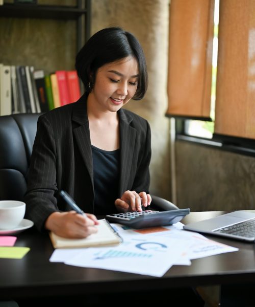 A beautiful and successful Asian businesswoman or female business entrepreneur calculates sales using a calculator and laptop computer at her desk in the office.