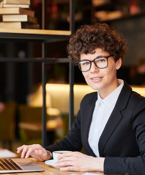 Young businesswoman in eyeglasses and suit sitting in front of camera by table, having tea and networking