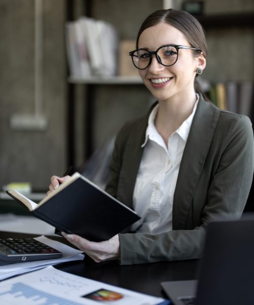 Young businesswoman smiling and holding notebook in modern office. Professional woman with glasses working in workplace with laptop and charts.
